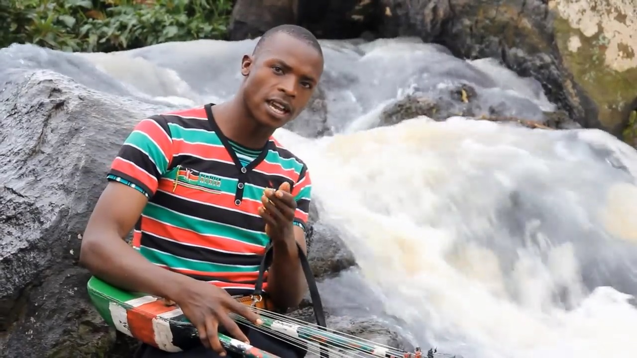 Amos Barasa in Bindu Bichenjanga video by a waterfall strumming litungu and gesturing foward
