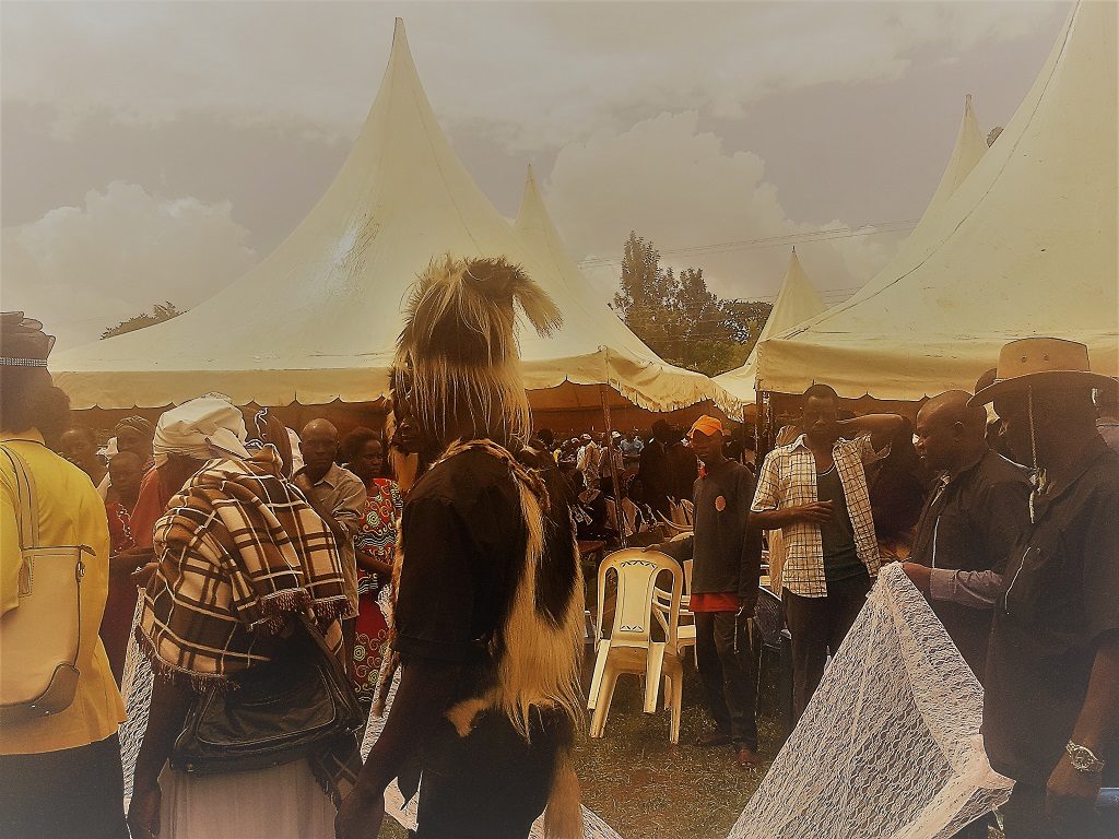 Image of four adults, one in traditional wear, in a procession holding white lace cloths during a burial ceremony as a crowd watches on