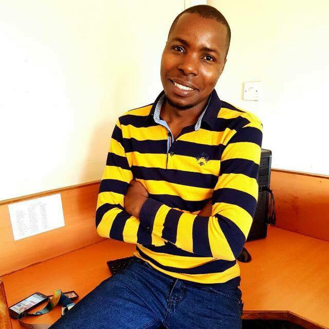 photo of Luhya comedy act  in yellow stripped polo shirt The musalia Mudambi smiling, arms folded leaning on table edge