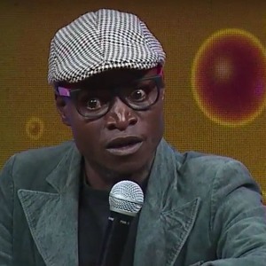 Luhya comedy stand up act Smart joker with blazer and golf hat on holding microphone 