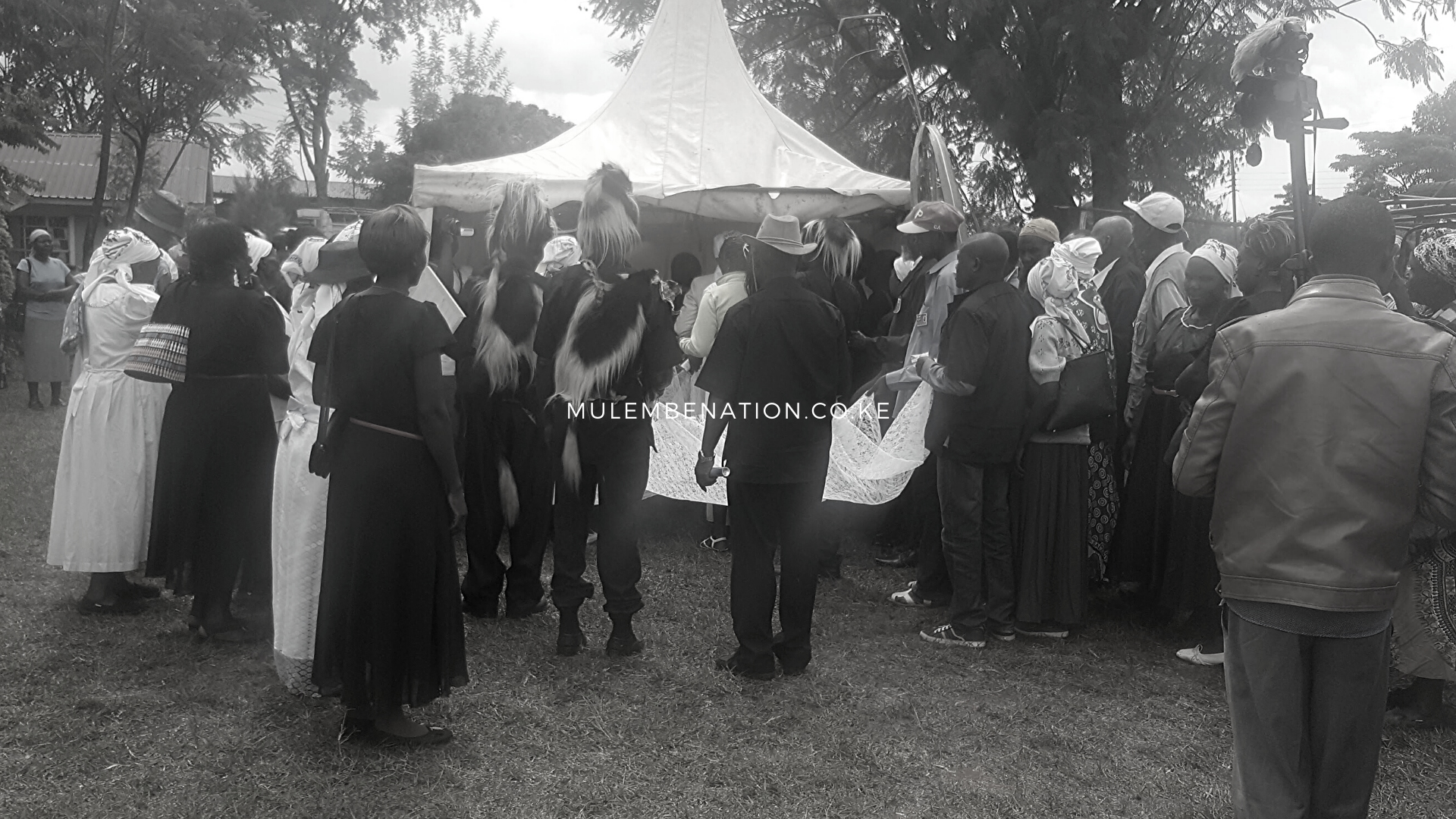Men and women, some in traditional attire, hold the ends of a lace material in preparation of draping the lace over a casket